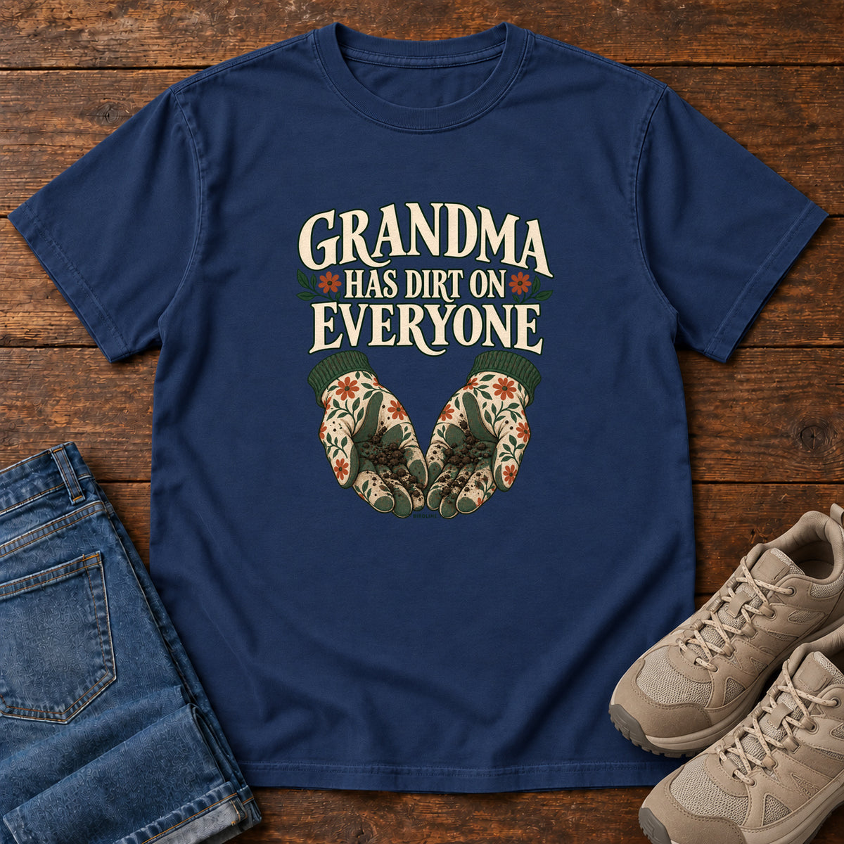 Grandma Has Dirt on Everyone T-Shirt in True Navy, front laid flat on wood, featuring floral gardening gloves cupping soil beneath large cream slogan lettering.