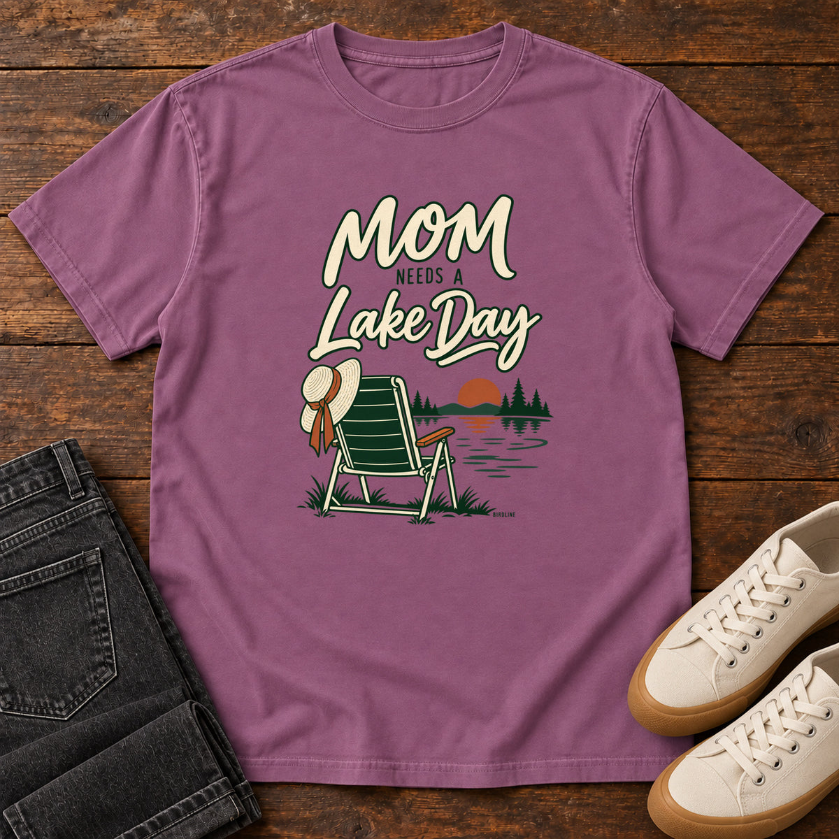 Berry Mom Needs a Lake Day T-Shirt with a striped folding chair, sunhat and lakeside sunset illustration, front flat lay on wood.