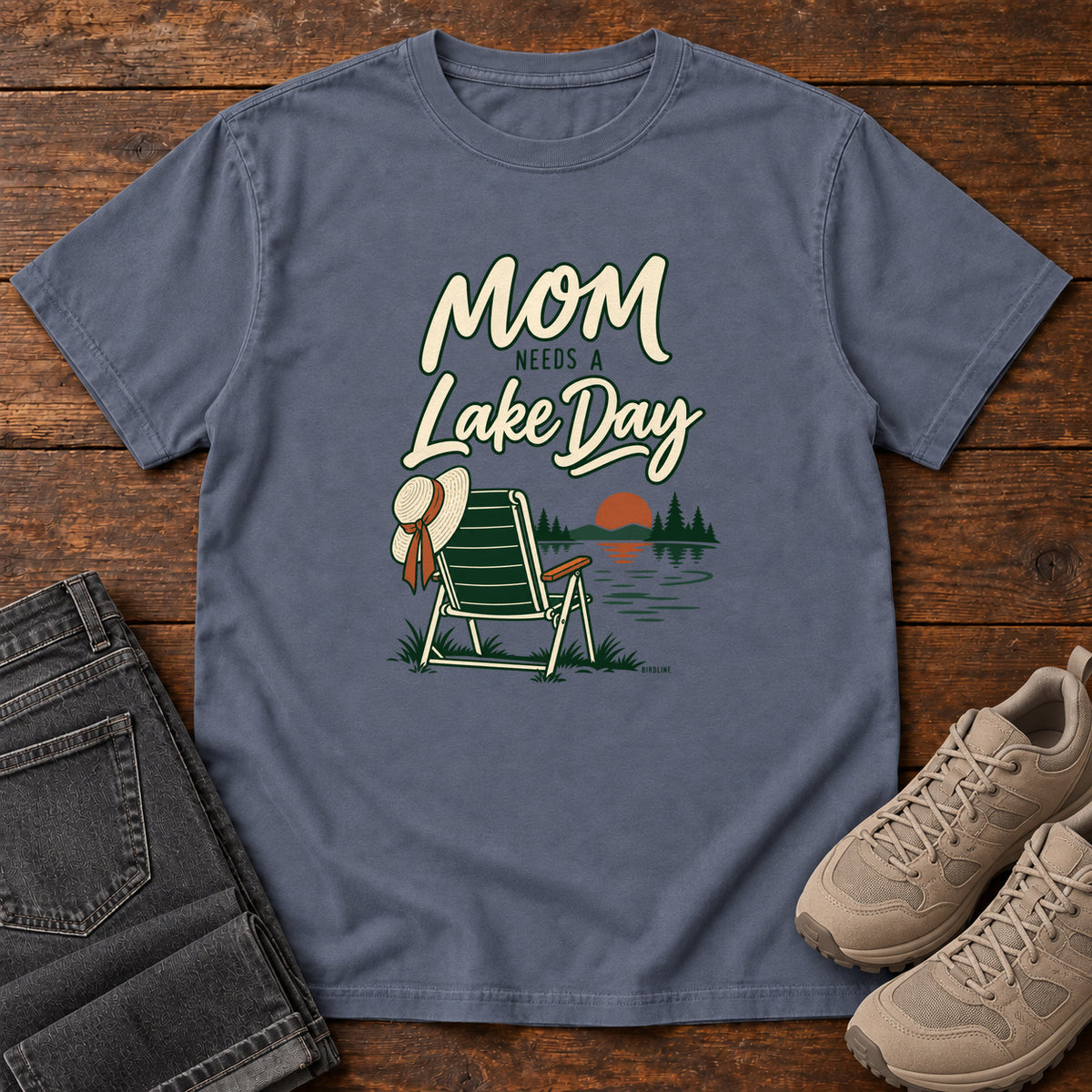 Denim Mom Needs a Lake Day T-Shirt with a striped folding chair, sunhat and lakeside sunset illustration, front flat lay on wood.
