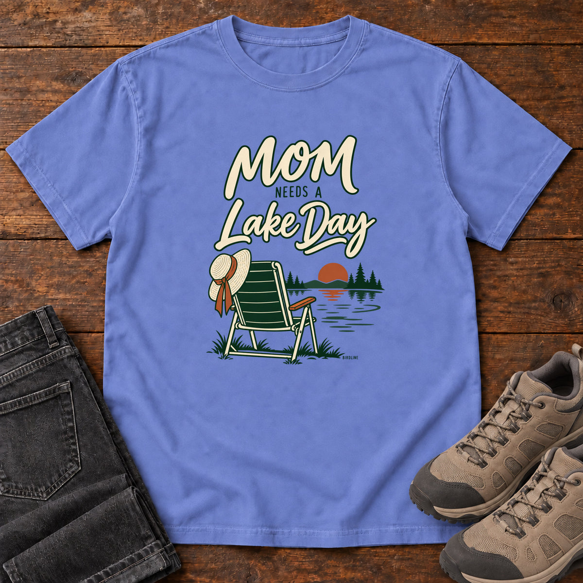 Flo Blue Mom Needs a Lake Day T-Shirt with a striped folding chair, sunhat and lakeside sunset illustration, front flat lay on wood.