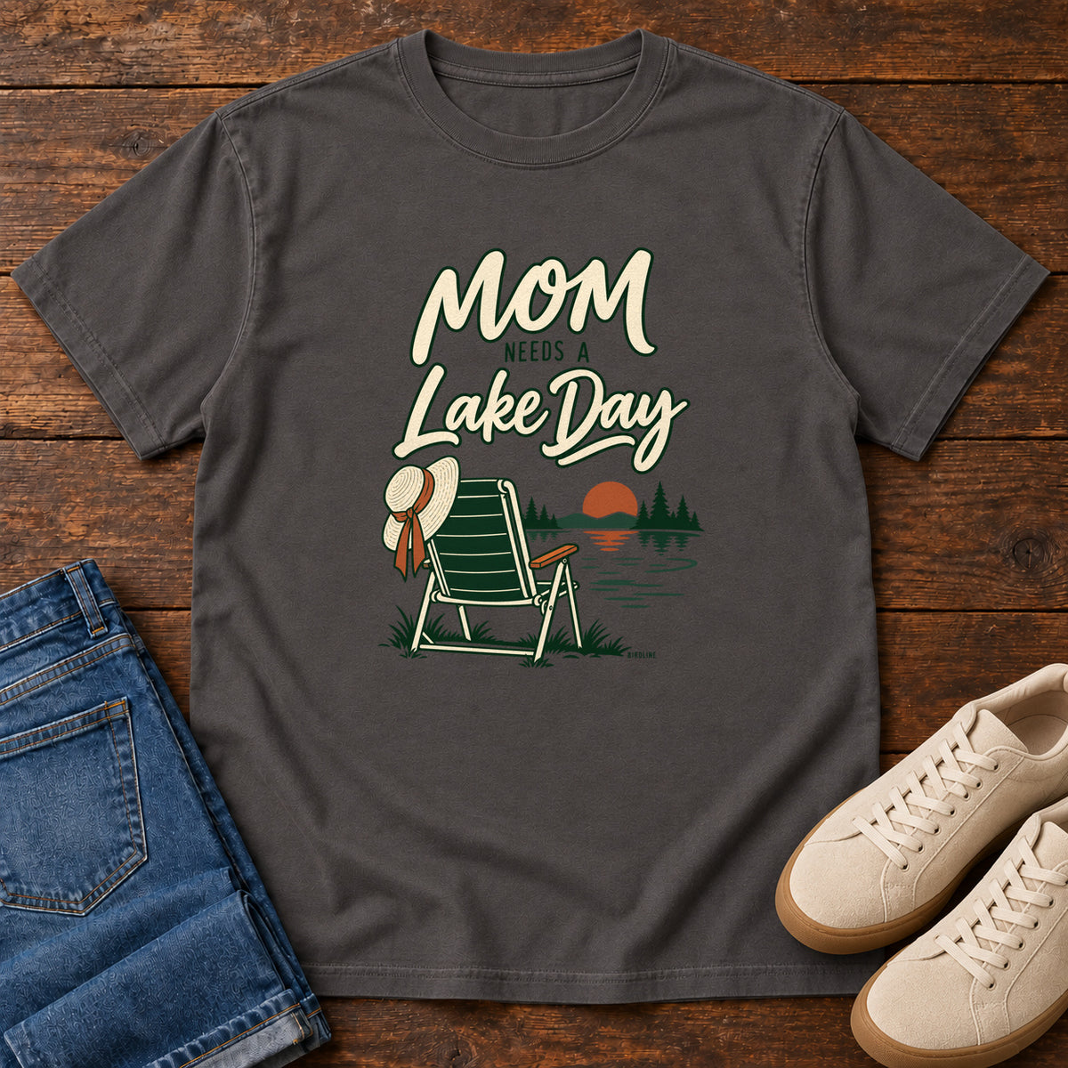 Graphite Mom Needs a Lake Day T-Shirt with a striped folding chair, sunhat and lakeside sunset illustration, front flat lay on wood.