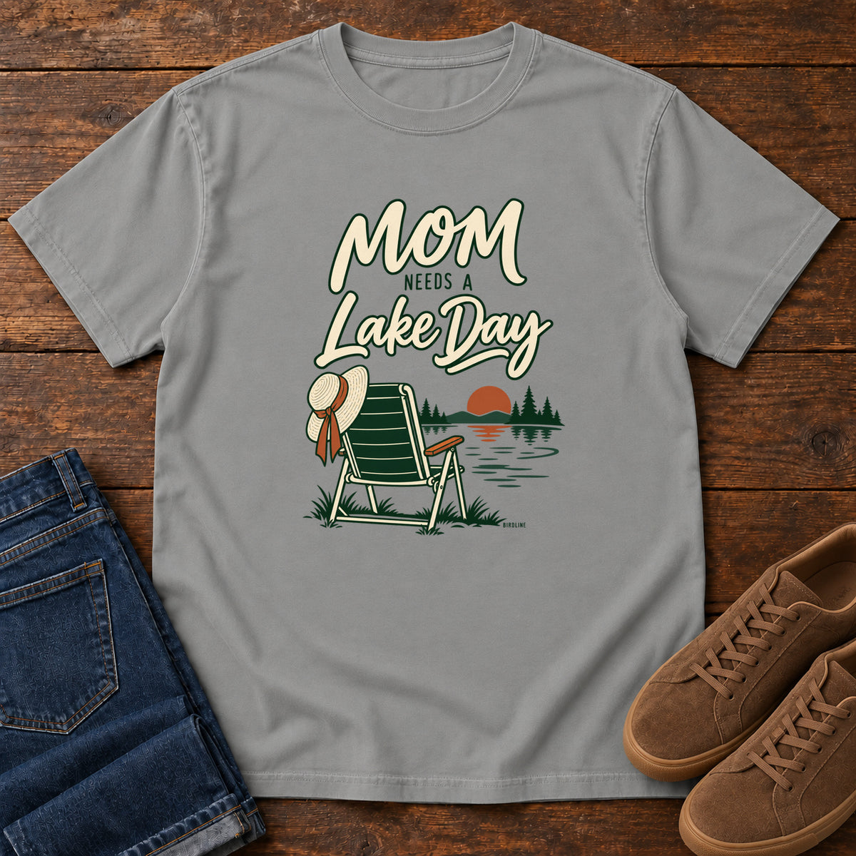 Grey Mom Needs a Lake Day T-Shirt with a striped folding chair, sunhat and lakeside sunset illustration, front flat lay on wood.