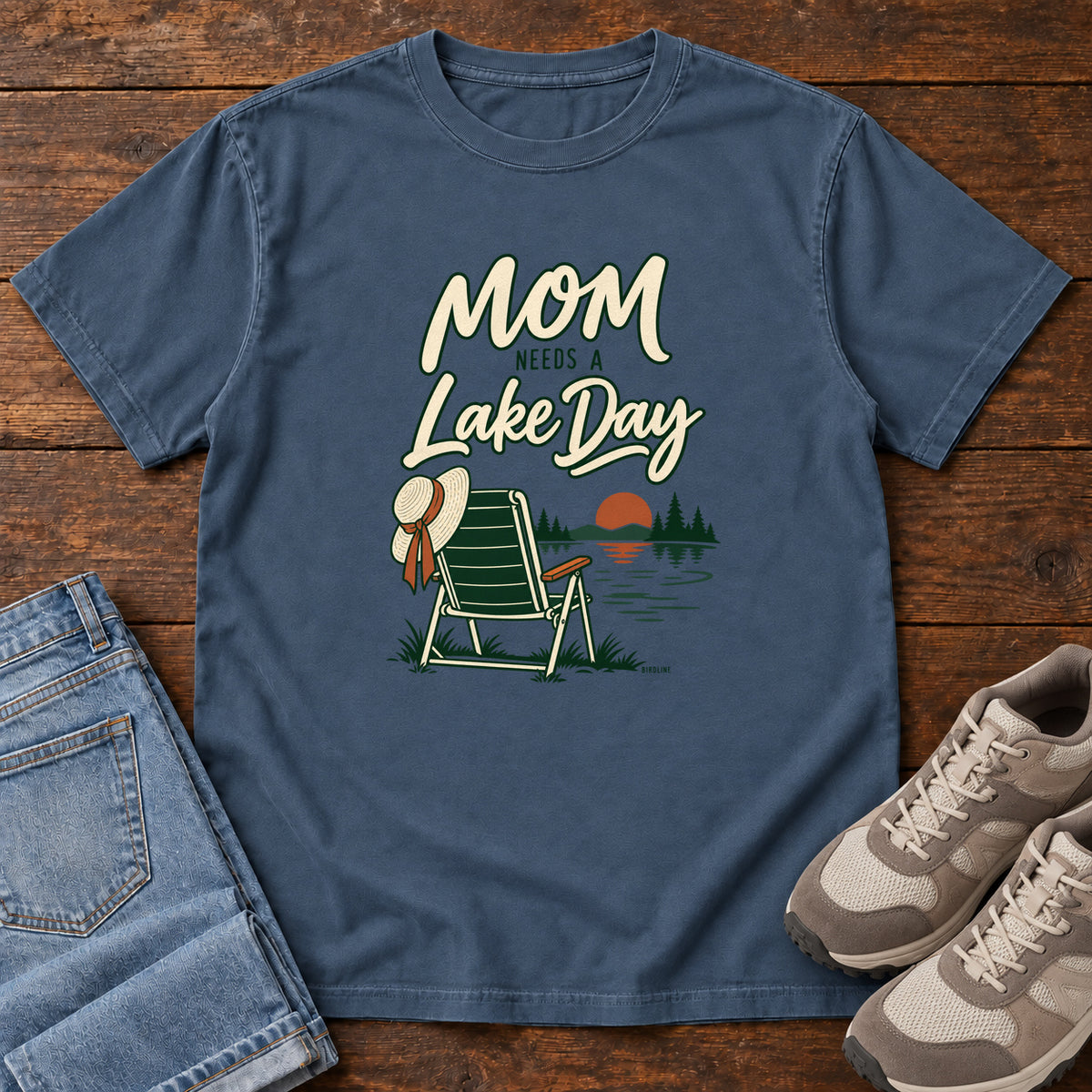 Midnight Mom Needs a Lake Day T-Shirt with a striped folding chair, sunhat and lakeside sunset illustration, front flat lay on wood.