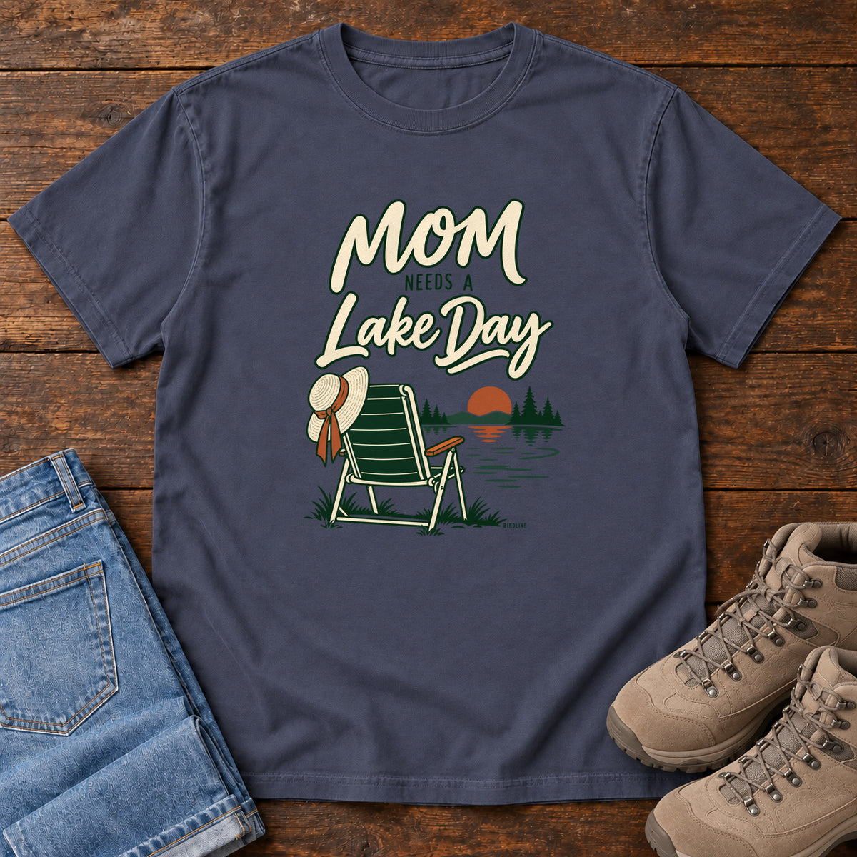 Navy Mom Needs a Lake Day T-Shirt with a striped folding chair, sunhat and lakeside sunset illustration, front flat lay on wood.