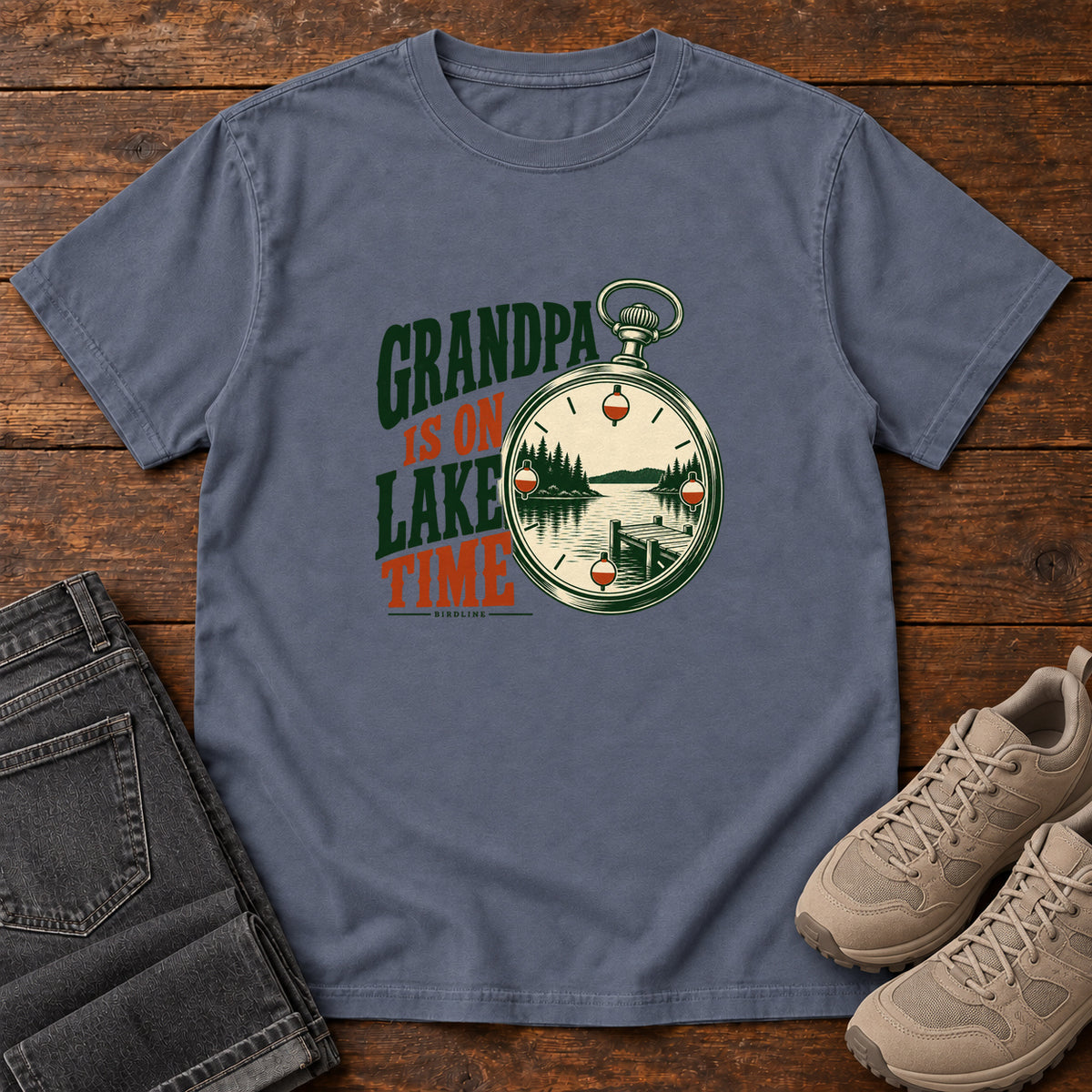 Denim Grandpa Is on Lake Time T-Shirt with a pocket watch framing a lake and dock beside green and orange lettering; front flat lay on wood.