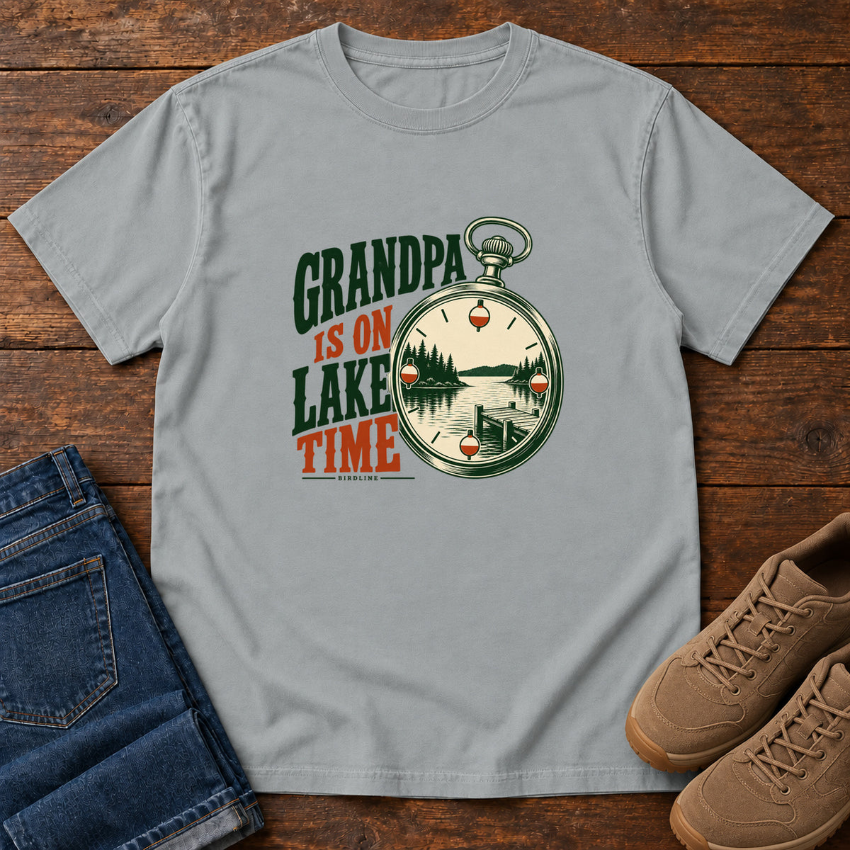 Granite Grandpa Is on Lake Time T-Shirt with a pocket watch framing a lake and dock beside green and orange lettering; front flat lay on wood.