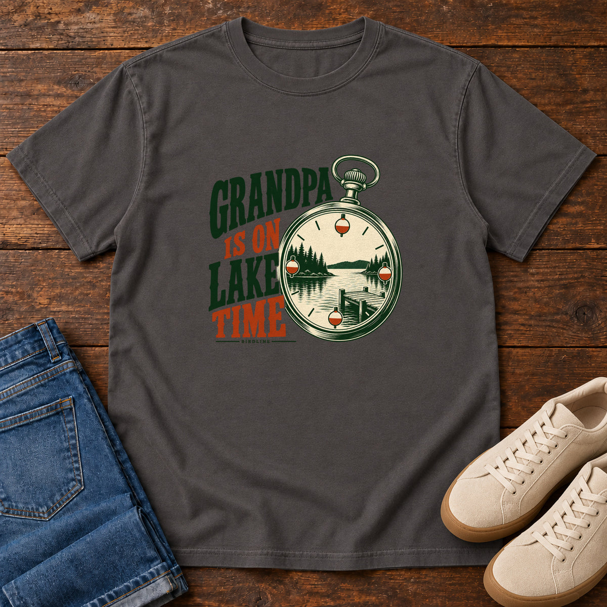 Graphite Grandpa Is on Lake Time T-Shirt with a pocket watch framing a lake and dock beside green and orange lettering; front flat lay on wood.