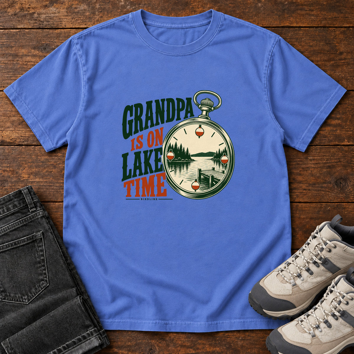 Mystic Blue Grandpa Is on Lake Time T-Shirt with a pocket watch framing a lake and dock beside green and orange lettering; front flat lay on wood.
