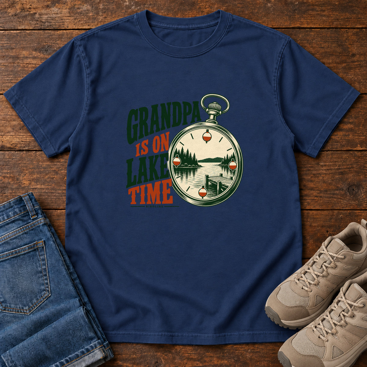 True Navy Grandpa Is on Lake Time T-Shirt with a pocket watch framing a lake and dock beside green and orange lettering; front flat lay on wood.