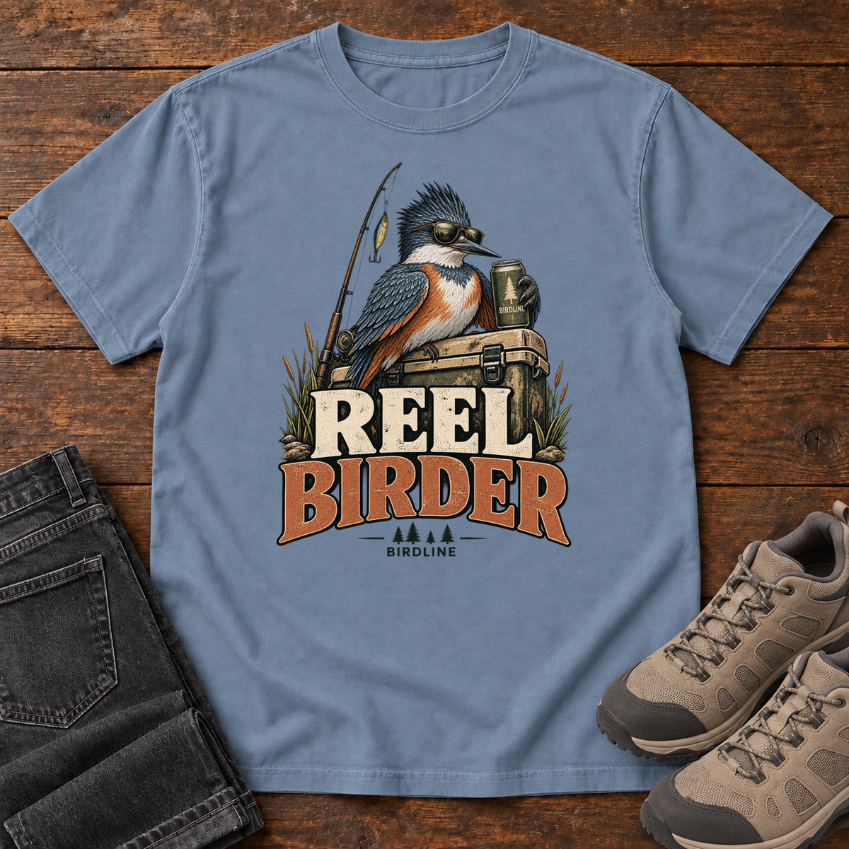Blue Jean Reel Birder T-Shirt with a crested bird in sunglasses holding a mug beside a fishing rod and tackle box; front flat lay on wood.