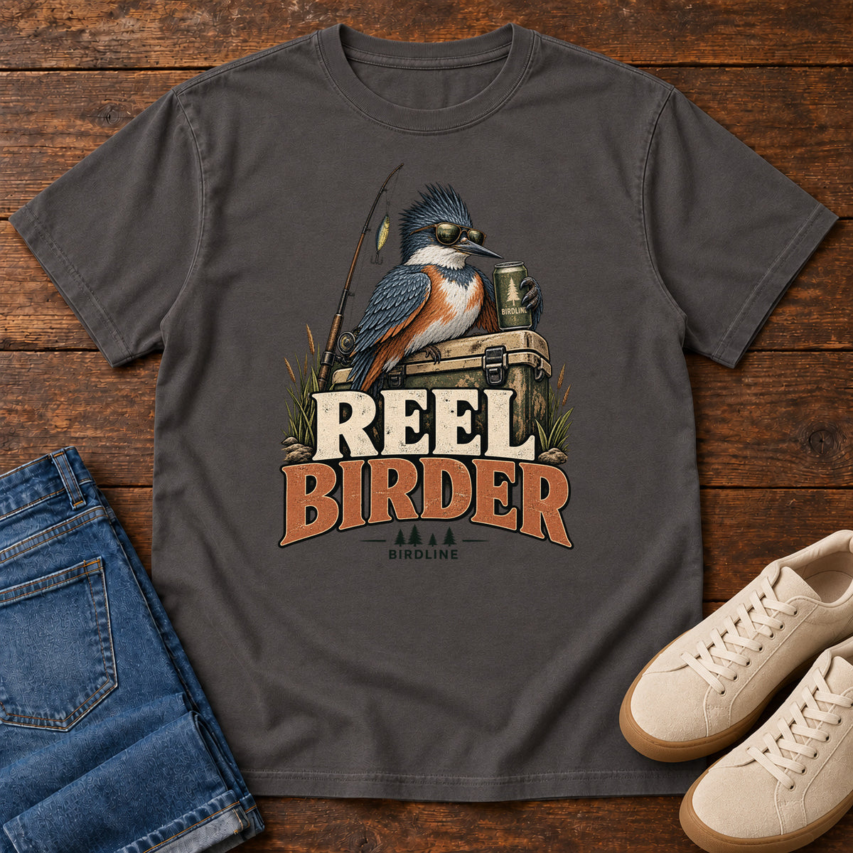 Graphite Reel Birder T-Shirt with a crested bird in sunglasses holding a mug beside a fishing rod and tackle box; front flat lay on wood.