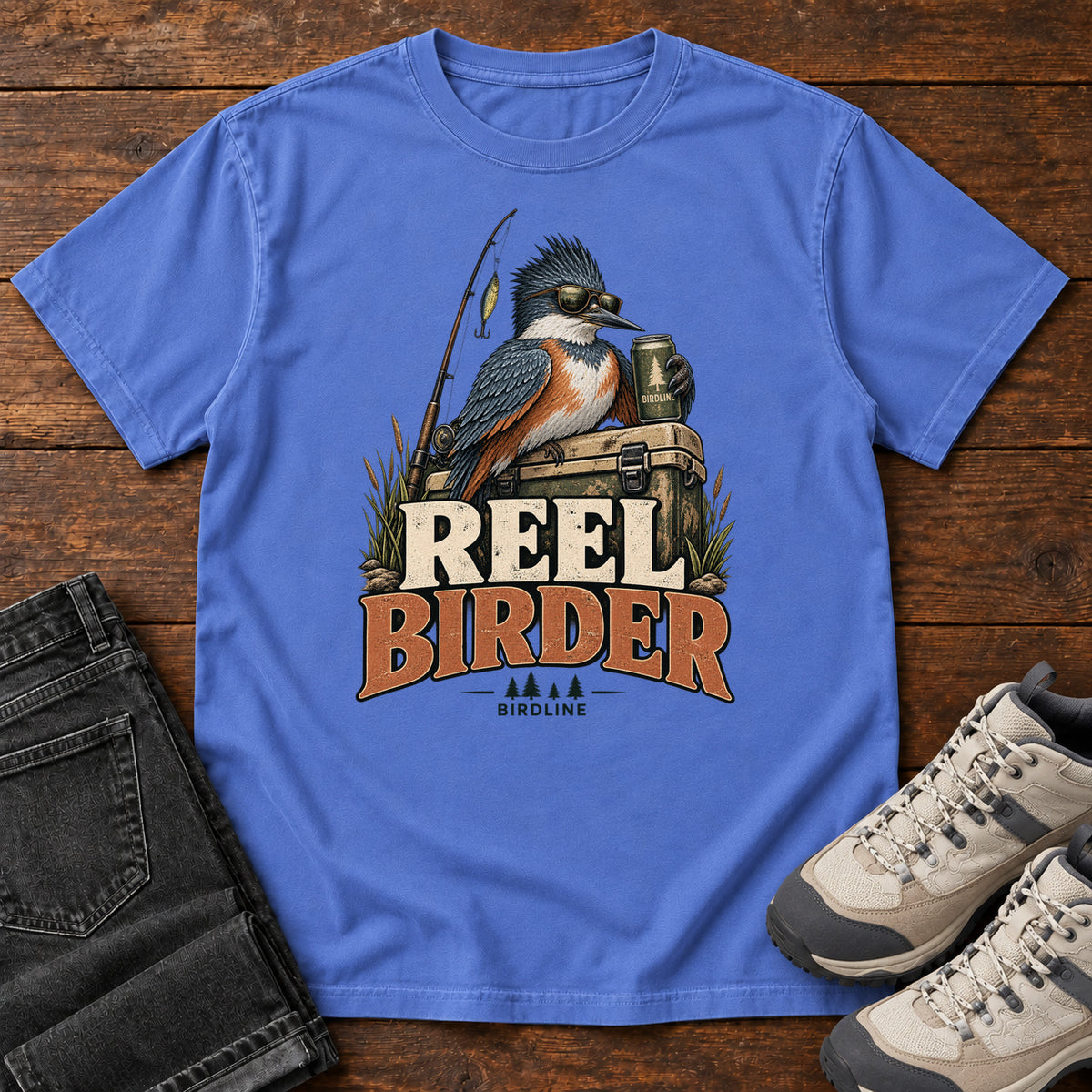 Mystic Blue Reel Birder T-Shirt with a crested bird in sunglasses holding a mug beside a fishing rod and tackle box; front flat lay on wood.