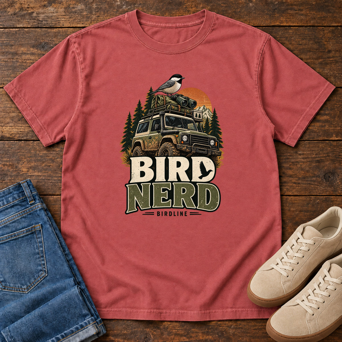 Crimson Bird Nerd Off-Roader T-Shirt with a small black-capped bird atop an off-road vehicle, trees and sunset above Bird Nerd lettering, front flat lay on wood.