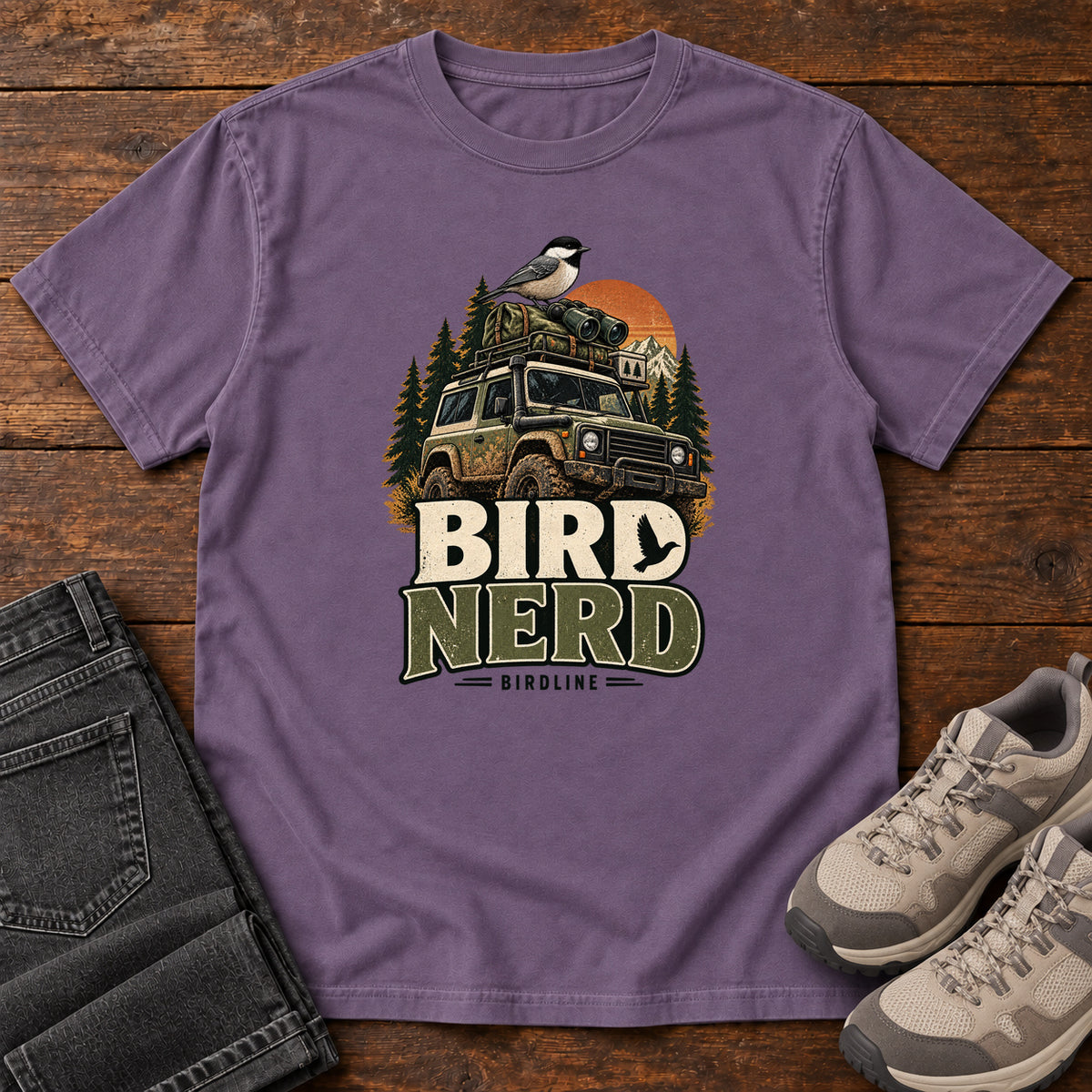Grape Bird Nerd Off-Roader T-Shirt with a small black-capped bird atop an off-road vehicle, trees and sunset above Bird Nerd lettering, front flat lay on wood.