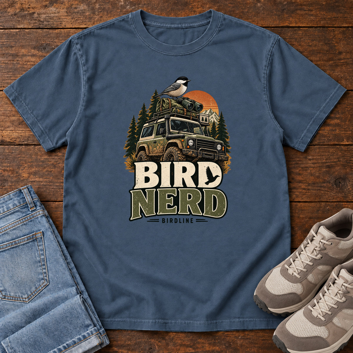 Midnight Bird Nerd Off-Roader T-Shirt with a small black-capped bird atop an off-road vehicle, trees and sunset above Bird Nerd lettering, front flat lay on wood.