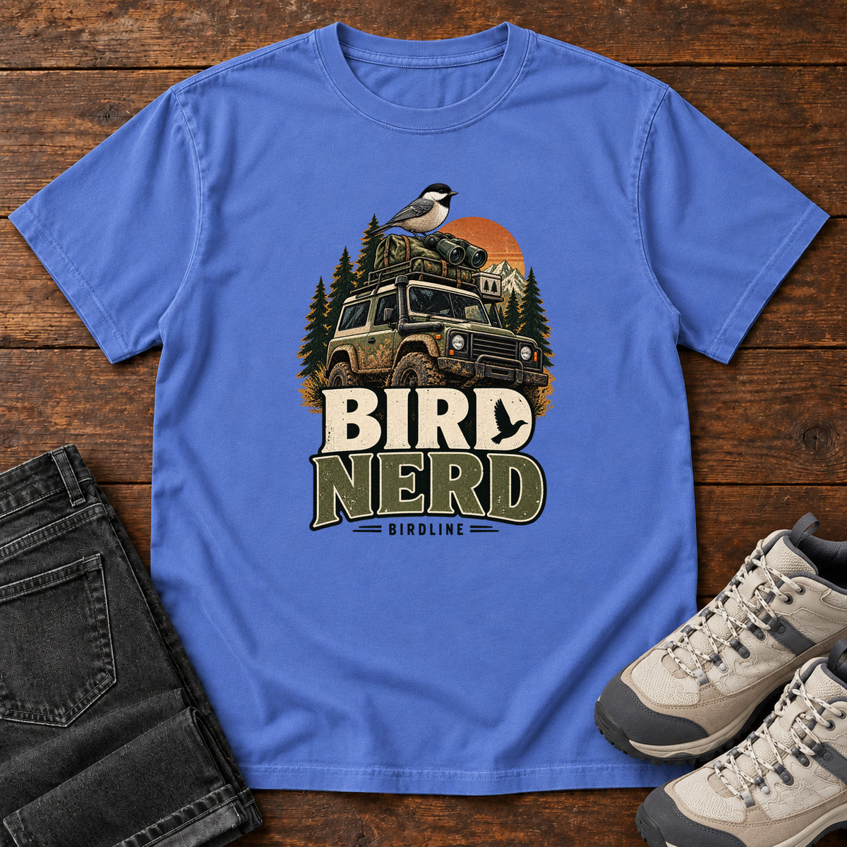 Mystic Blue Bird Nerd Off-Roader T-Shirt with a small black-capped bird atop an off-road vehicle, trees and sunset above Bird Nerd lettering, front flat lay on wood.