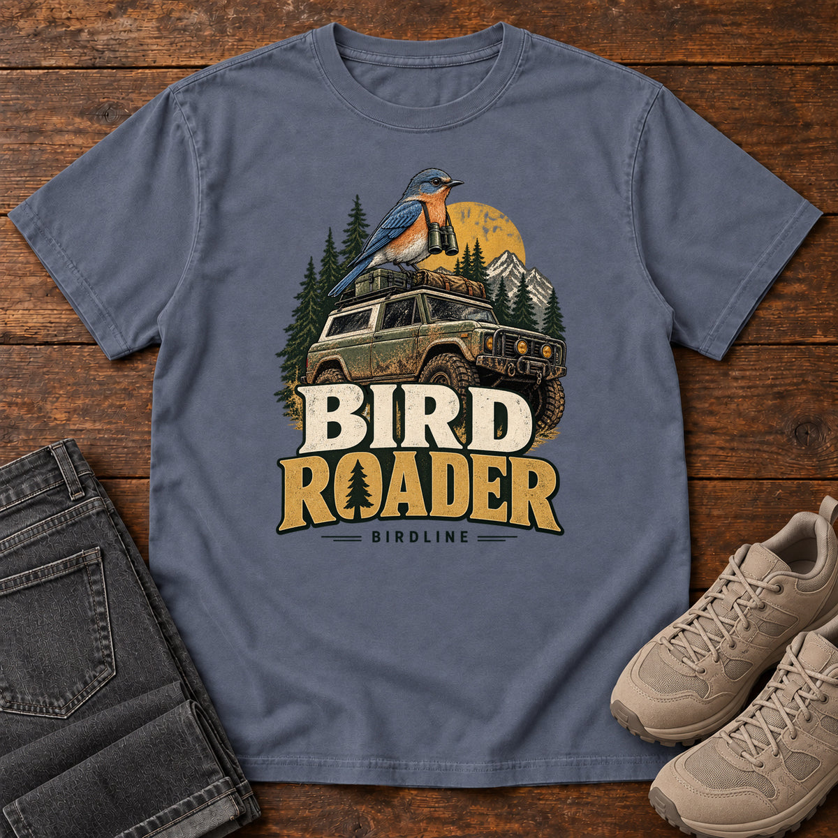 Bird Roader T-Shirt in Denim, front laid flat on wood, featuring a blue-and-orange bird above an off-road vehicle, pine trees, mountains and a yellow sun.
