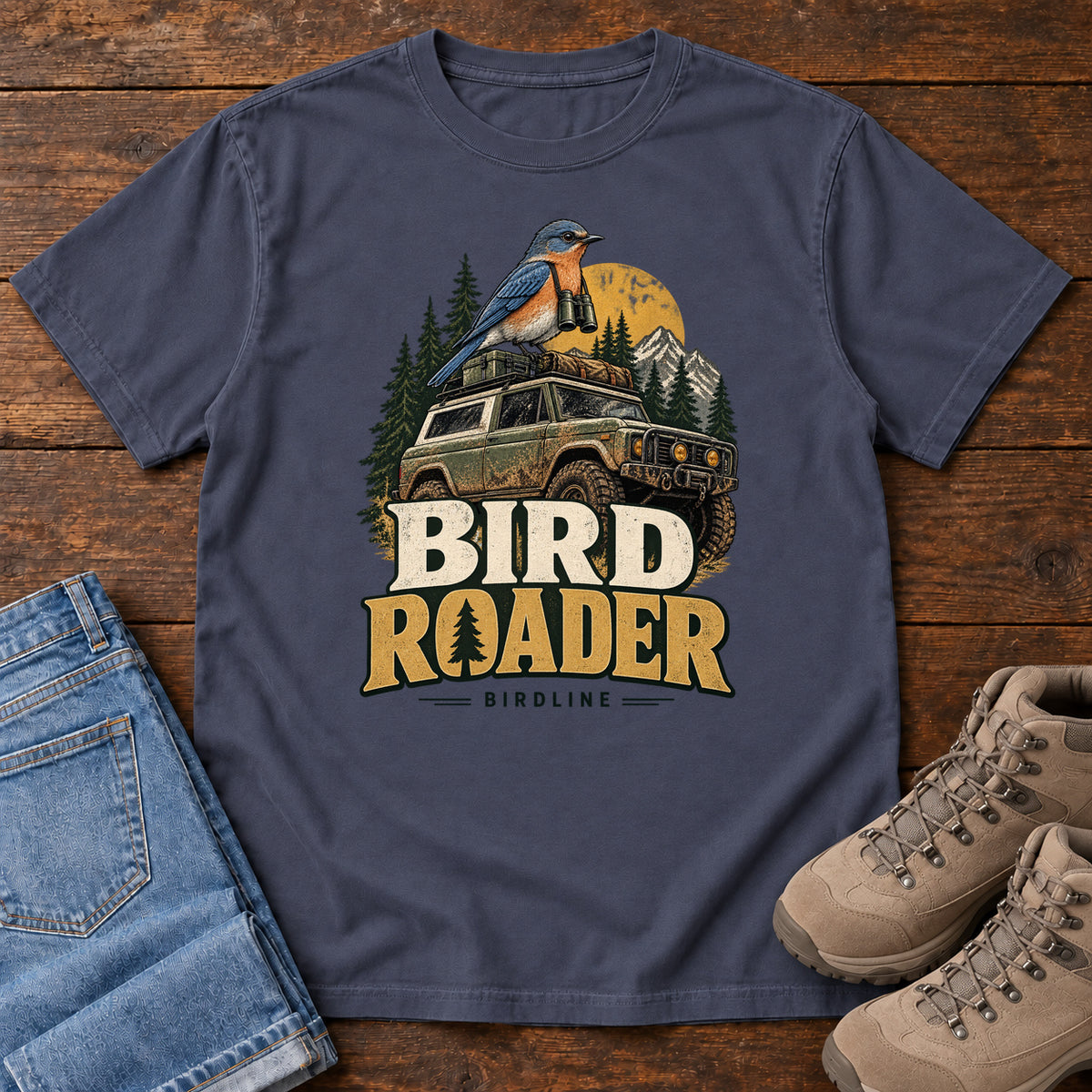 Bird Roader T-Shirt in Navy, front laid flat on wood, featuring a blue-and-orange bird above an off-road vehicle, pine trees, mountains and a yellow sun.
