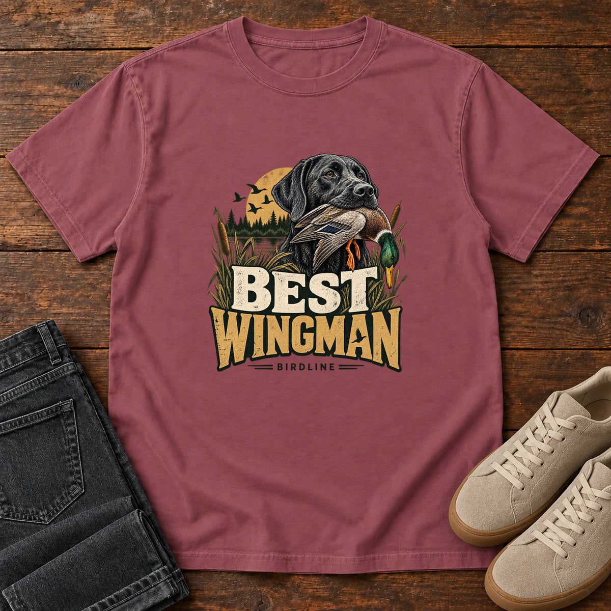 Brick Best Wingman T-Shirt with a black dog carrying a duck, marsh reeds and cream-and-gold lettering, front flat lay on wood.