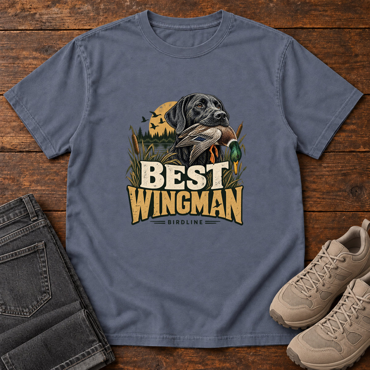 Denim Best Wingman T-Shirt with a black dog carrying a duck, marsh reeds and cream-and-gold lettering, front flat lay on wood.