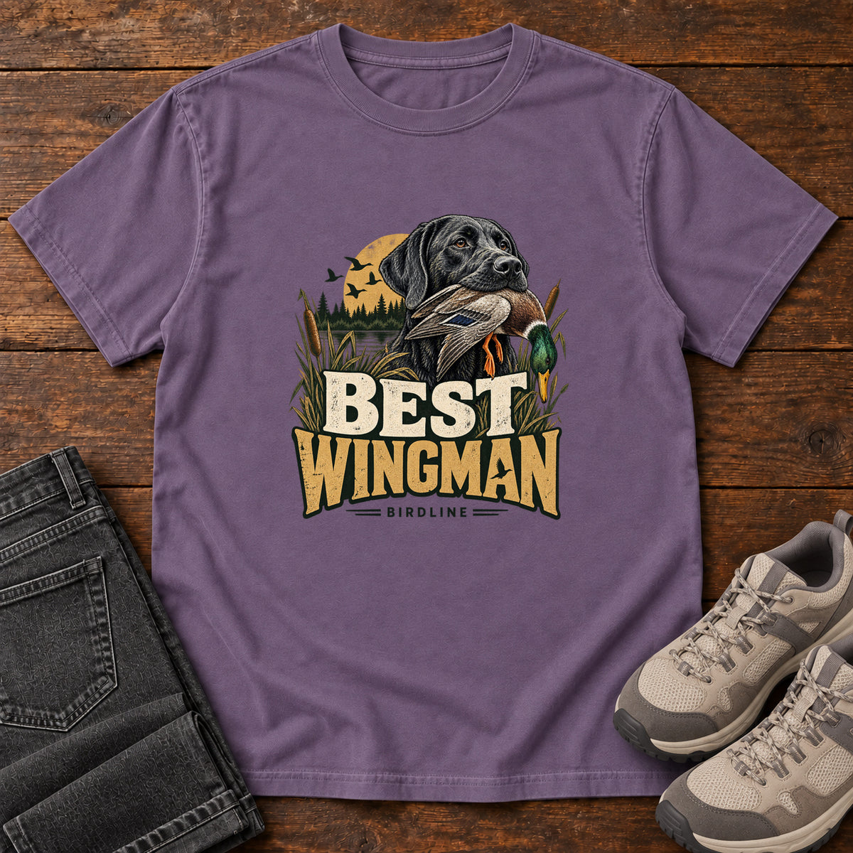 Grape Best Wingman T-Shirt with a black dog carrying a duck, marsh reeds and cream-and-gold lettering, front flat lay on wood.