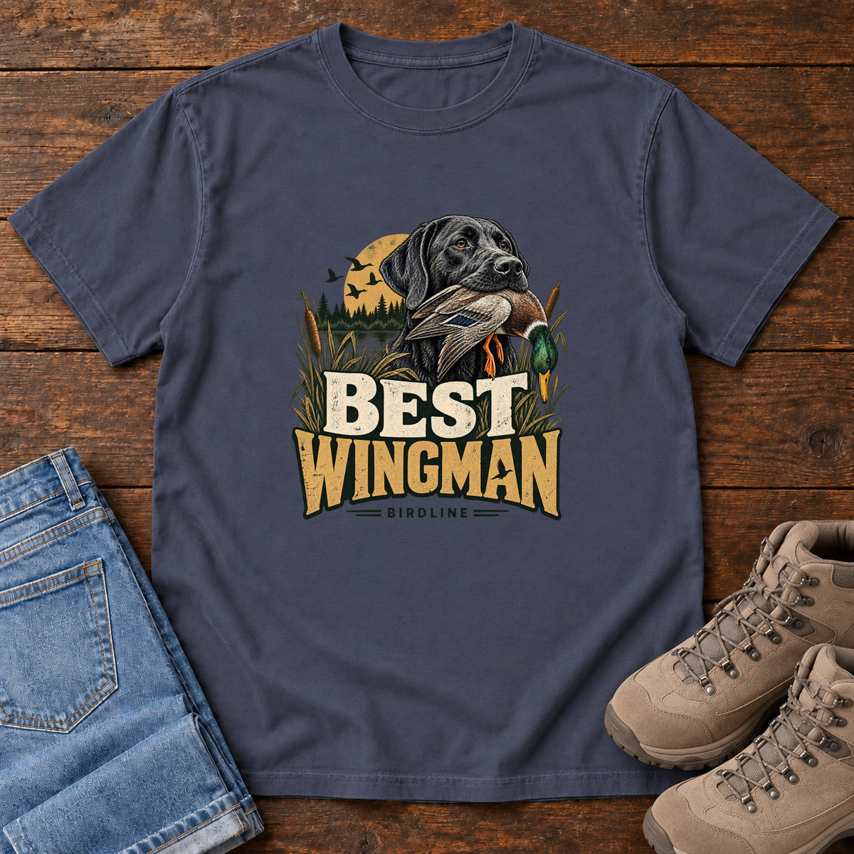 Navy Best Wingman T-Shirt with a black dog carrying a duck, marsh reeds and cream-and-gold lettering, front flat lay on wood.