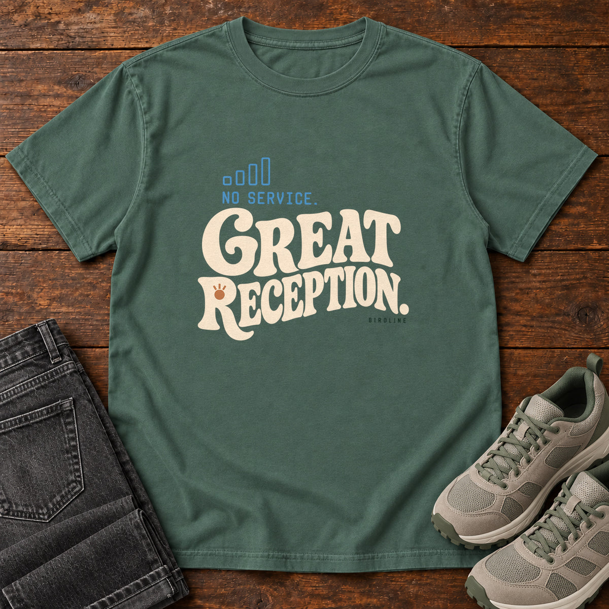 Blue Spruce No Service. Great Reception. T-Shirt with cream retro lettering and blue outlined phone-signal bars; front flat lay on wood.