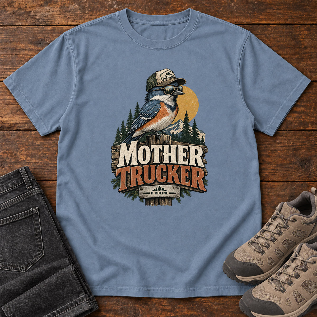 Blue Jean Mother Trucker T-Shirt with a blue-and-orange bird wearing a cap and sunglasses above mountains and pines; front flat lay on wood.