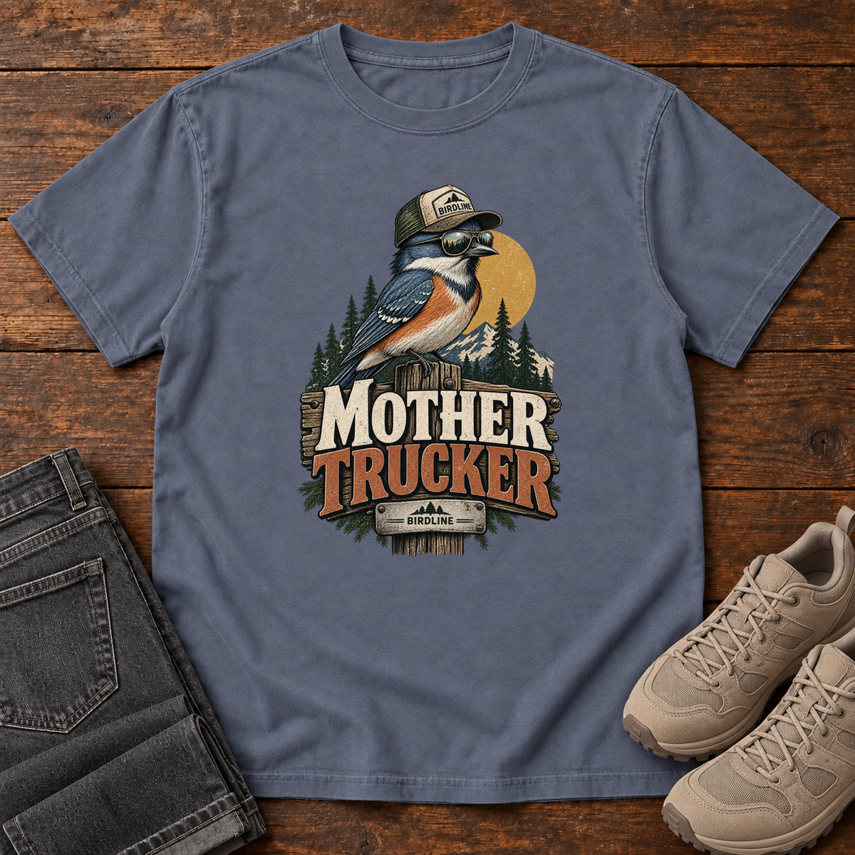 Denim Mother Trucker T-Shirt with a blue-and-orange bird wearing a cap and sunglasses above mountains and pines; front flat lay on wood.