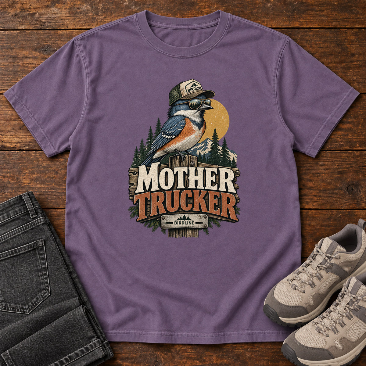Grape Mother Trucker T-Shirt with a blue-and-orange bird wearing a cap and sunglasses above mountains and pines; front flat lay on wood.