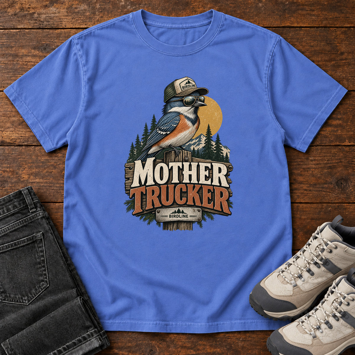 Mystic Blue Mother Trucker T-Shirt with a blue-and-orange bird wearing a cap and sunglasses above mountains and pines; front flat lay on wood.