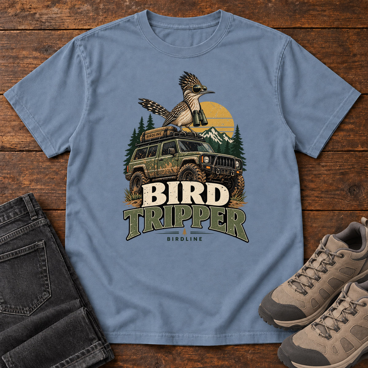 Blue Jean Bird Tripper T-Shirt with a crested, long-tailed bird with binoculars above an off-road vehicle and Bird Tripper lettering, front flat lay on wood.