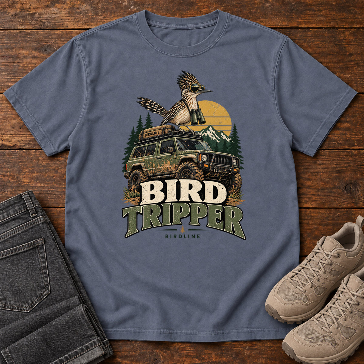 Denim Bird Tripper T-Shirt with a crested, long-tailed bird with binoculars above an off-road vehicle and Bird Tripper lettering, front flat lay on wood.