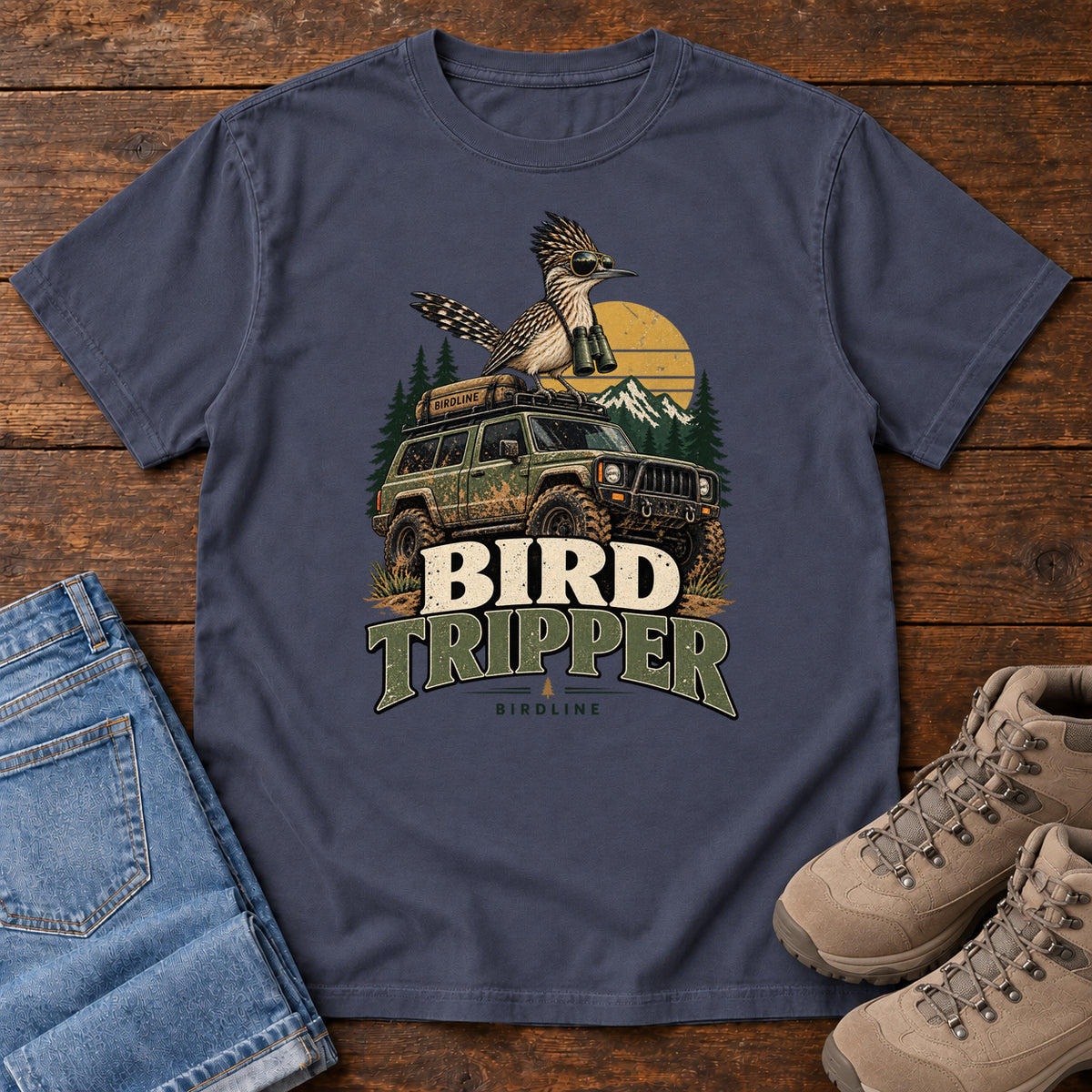 Navy Bird Tripper T-Shirt with a crested, long-tailed bird with binoculars above an off-road vehicle and Bird Tripper lettering, front flat lay on wood.