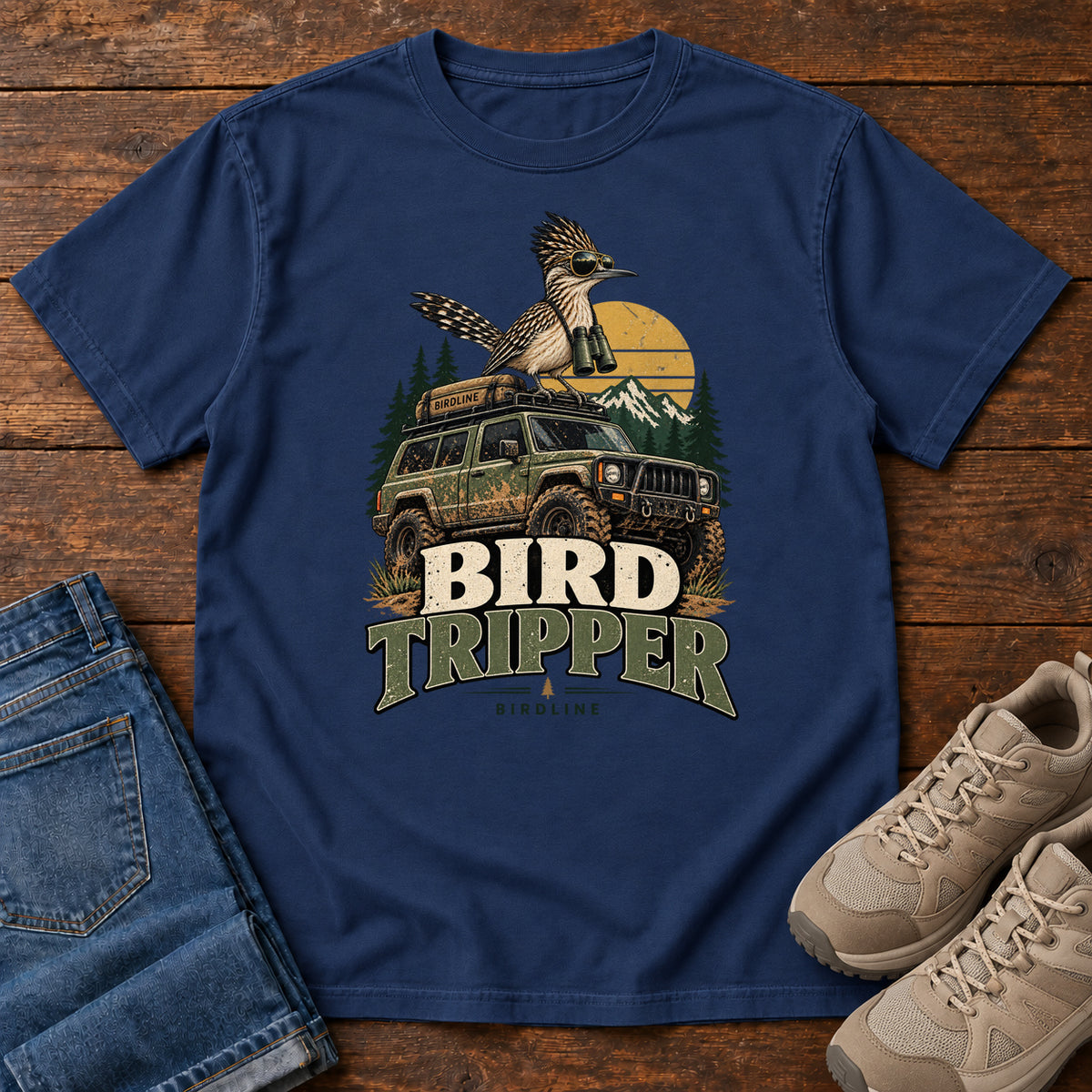 True Navy Bird Tripper T-Shirt with a crested, long-tailed bird with binoculars above an off-road vehicle and Bird Tripper lettering, front flat lay on wood.