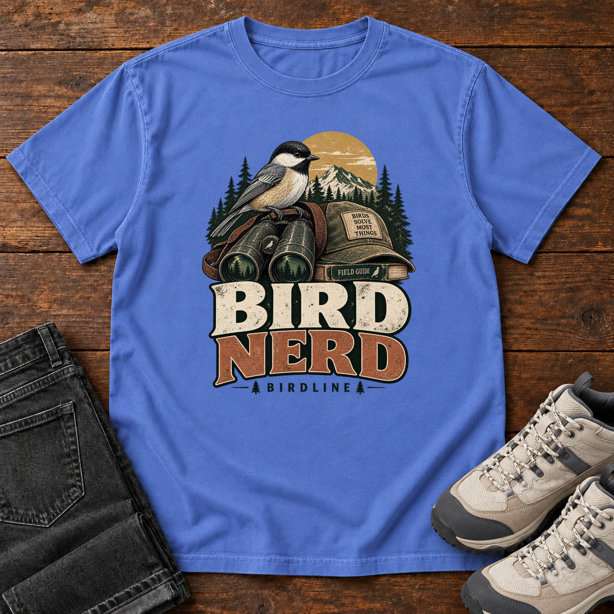 Bird Nerd Binoculars T-Shirt in Mystic Blue, front laid flat on wood, featuring a black-capped, cream-and-gray bird above binoculars and a cap, with mountains and bold lettering.