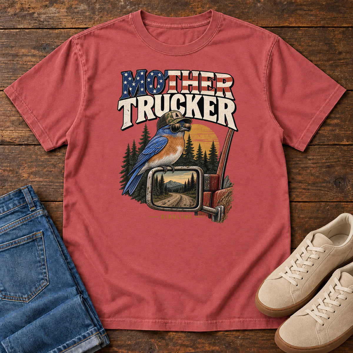 Crimson Mother Trucker Side Mirror T-Shirt with a blue-and-orange bird perched on a truck mirror beneath flag-patterned lettering, front flat lay on wood.