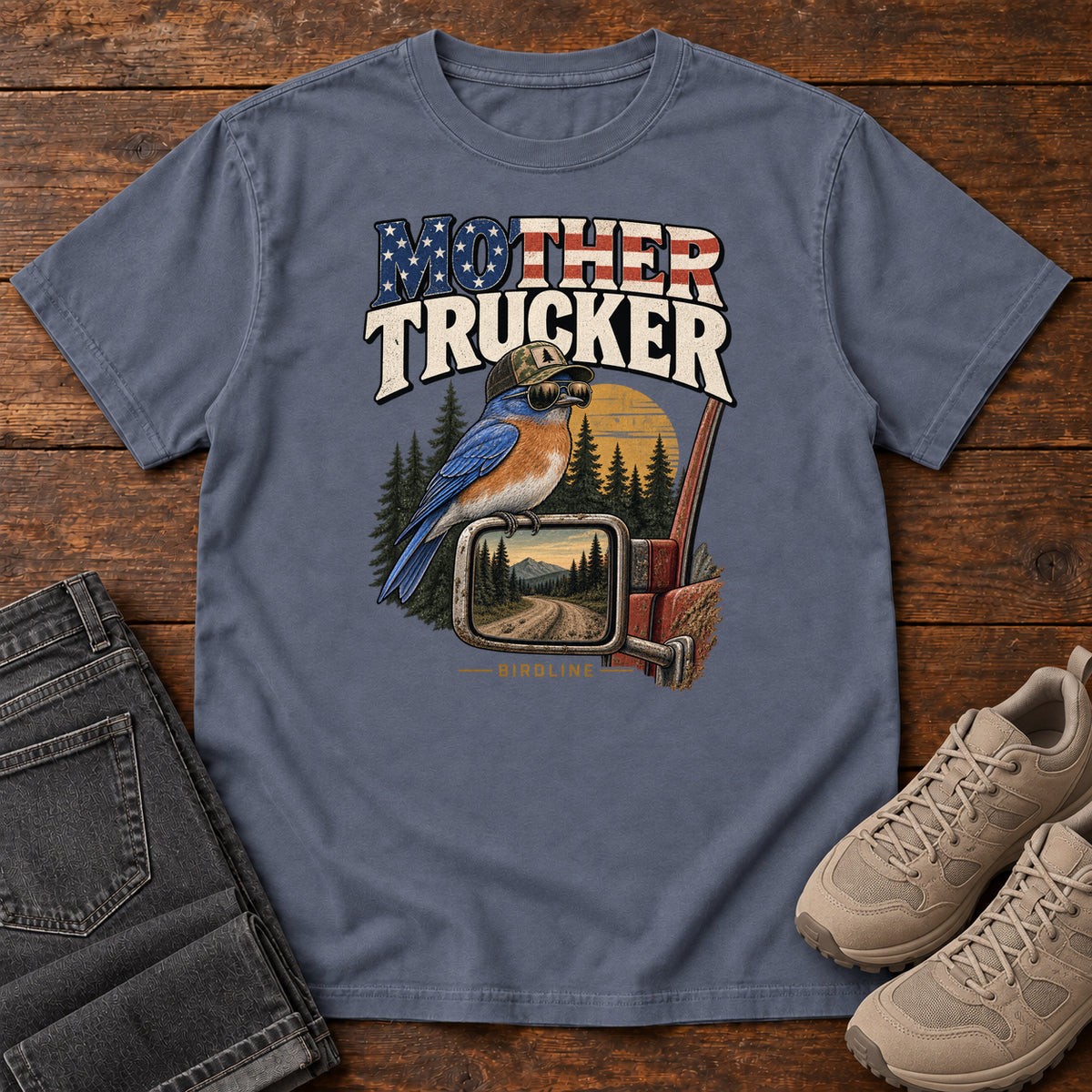 Denim Mother Trucker Side Mirror T-Shirt with a blue-and-orange bird perched on a truck mirror beneath flag-patterned lettering, front flat lay on wood.