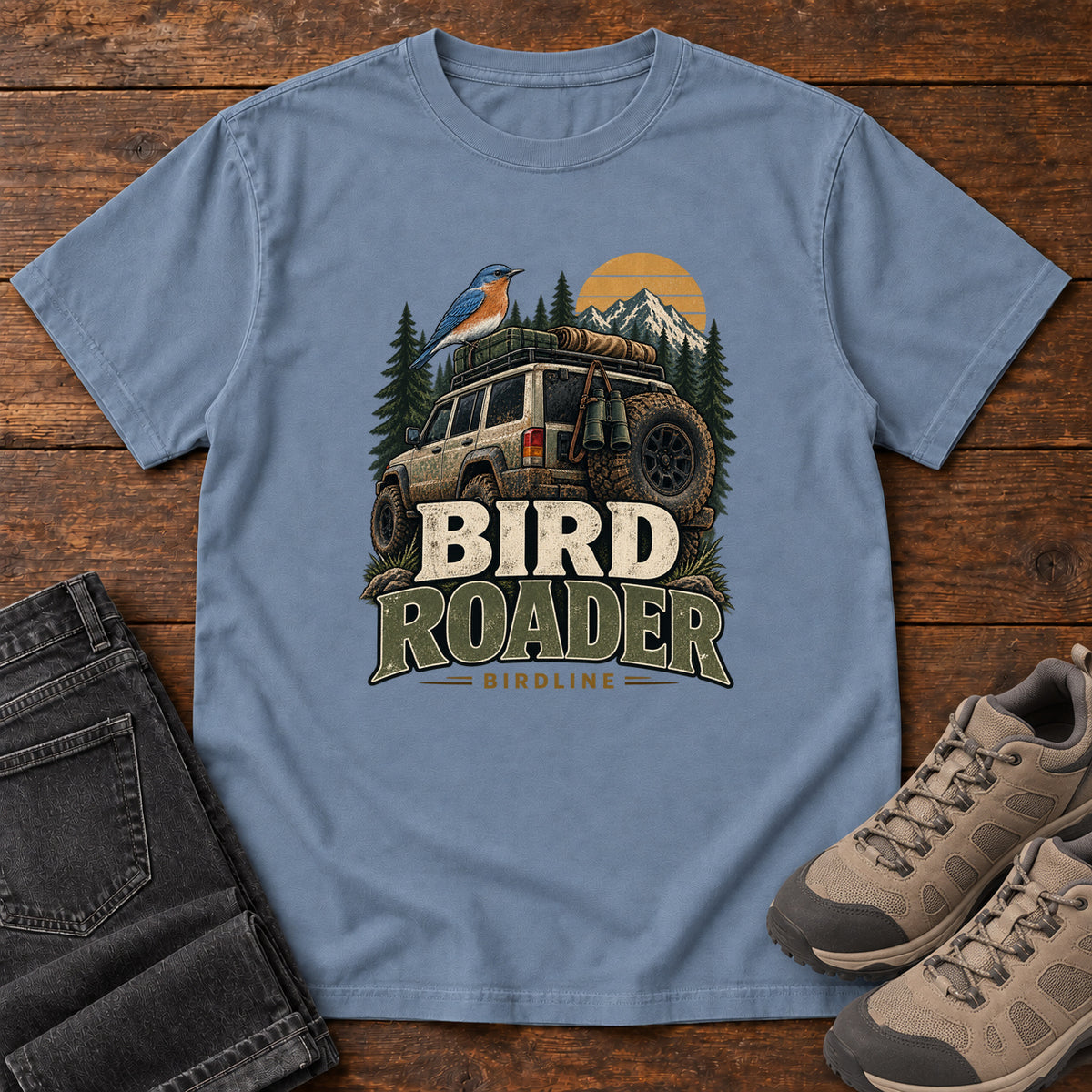Blue Jean Bird Roader Tailgate T-Shirt with a bird on an off-road vehicle beneath mountains and a golden sun; front flat lay on wood.