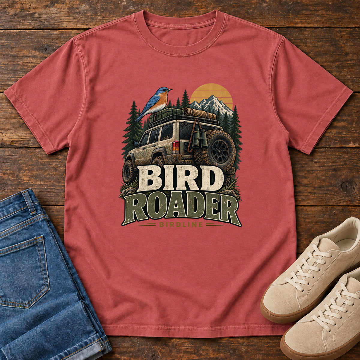 Crimson Bird Roader Tailgate T-Shirt with a bird on an off-road vehicle beneath mountains and a golden sun; front flat lay on wood.