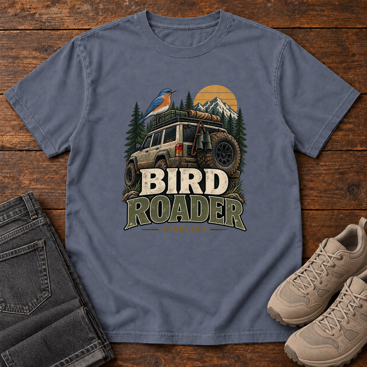Denim Bird Roader Tailgate T-Shirt with a bird on an off-road vehicle beneath mountains and a golden sun; front flat lay on wood.