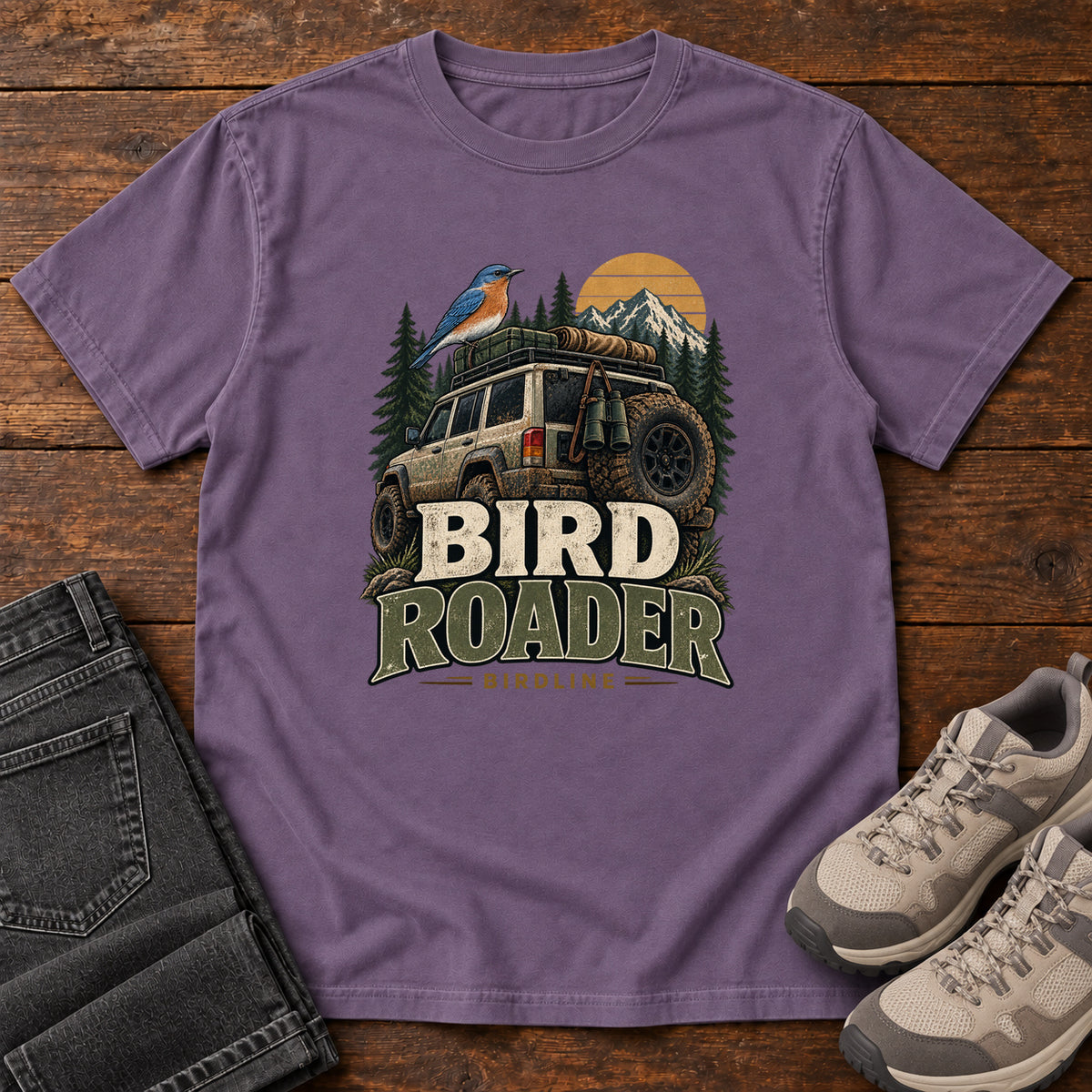 Grape Bird Roader Tailgate T-Shirt with a bird on an off-road vehicle beneath mountains and a golden sun; front flat lay on wood.