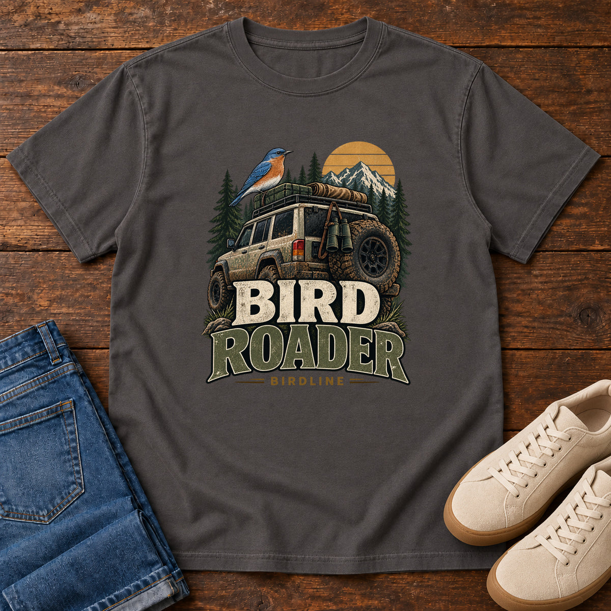 Graphite Bird Roader Tailgate T-Shirt with a bird on an off-road vehicle beneath mountains and a golden sun; front flat lay on wood.