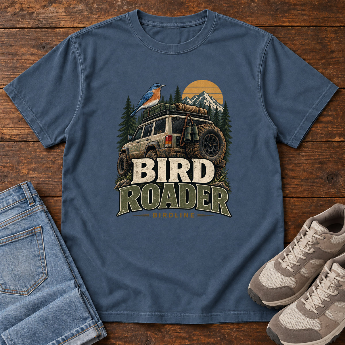 Midnight Bird Roader Tailgate T-Shirt with a bird on an off-road vehicle beneath mountains and a golden sun; front flat lay on wood.