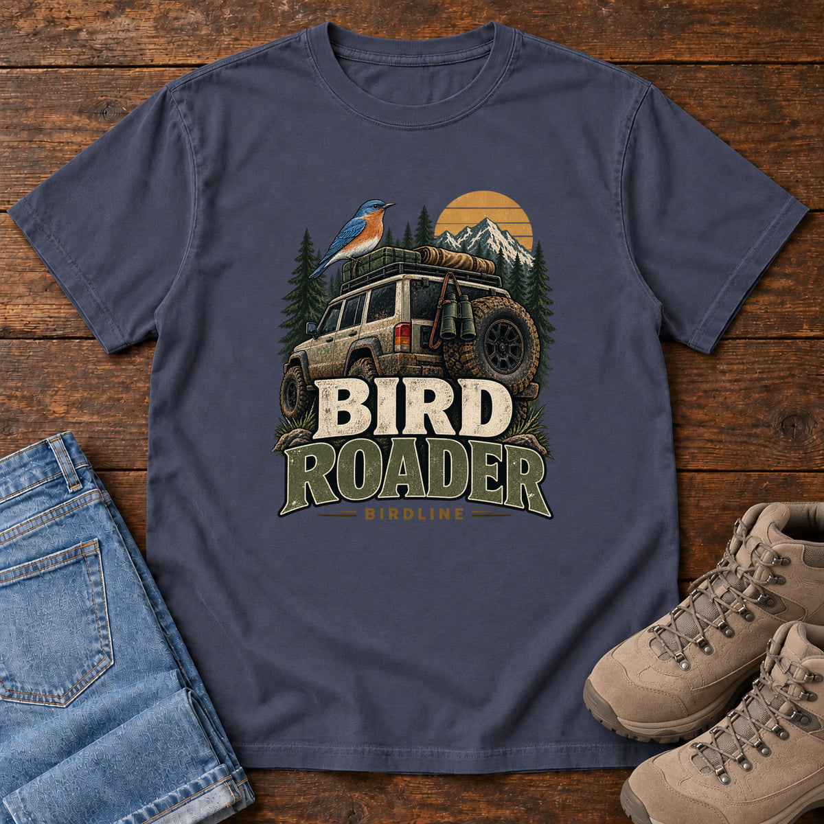 Navy Bird Roader Tailgate T-Shirt with a bird on an off-road vehicle beneath mountains and a golden sun; front flat lay on wood.