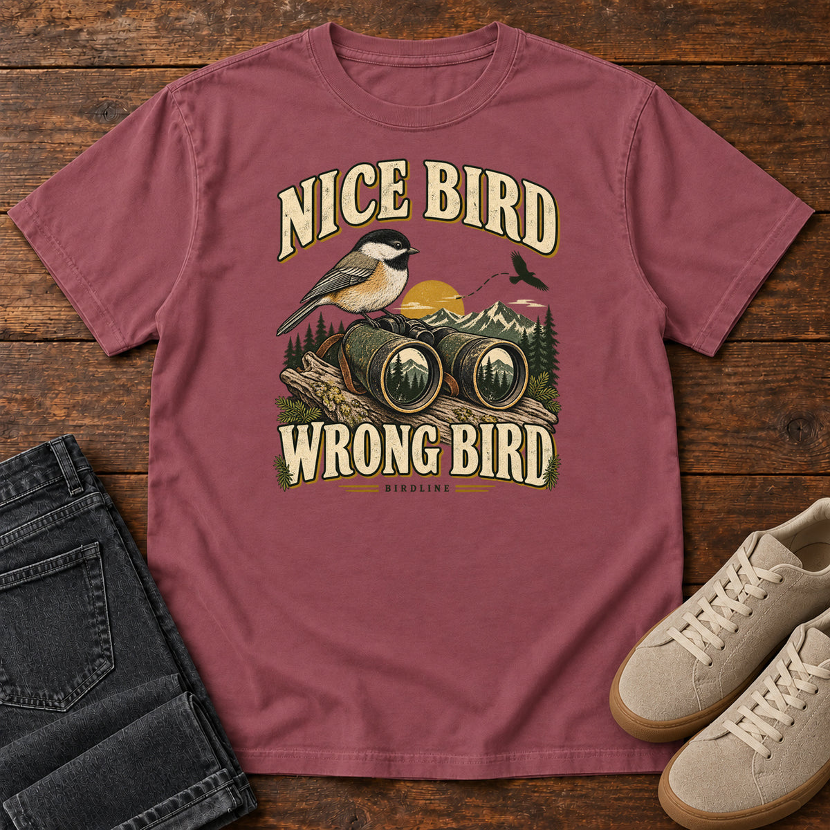 Brick Nice Bird. Wrong Bird. T-Shirt with a small black-capped bird on binoculars against mountains and a golden sun; front flat lay on wood.