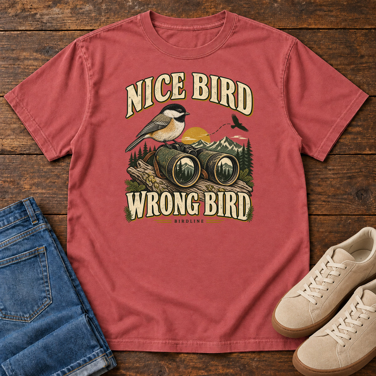 Crimson Nice Bird. Wrong Bird. T-Shirt with a small black-capped bird on binoculars against mountains and a golden sun; front flat lay on wood.