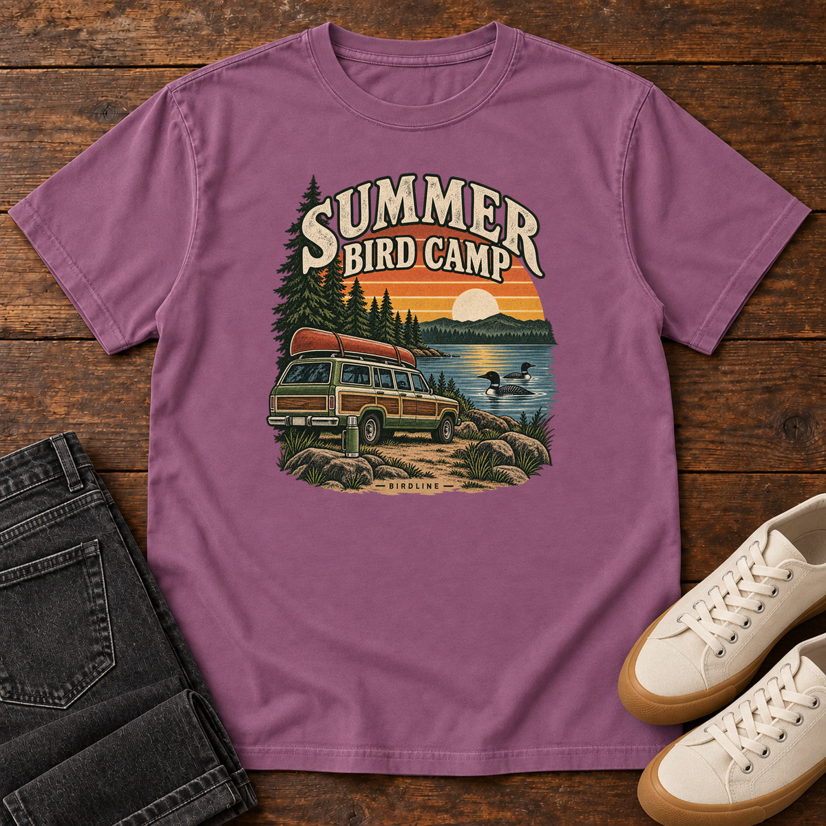 Summer Bird Camp T-Shirt in Berry, front laid flat on wood, featuring a canoe-topped vehicle beside a lake with swimming birds, pine trees and a striped sunset.