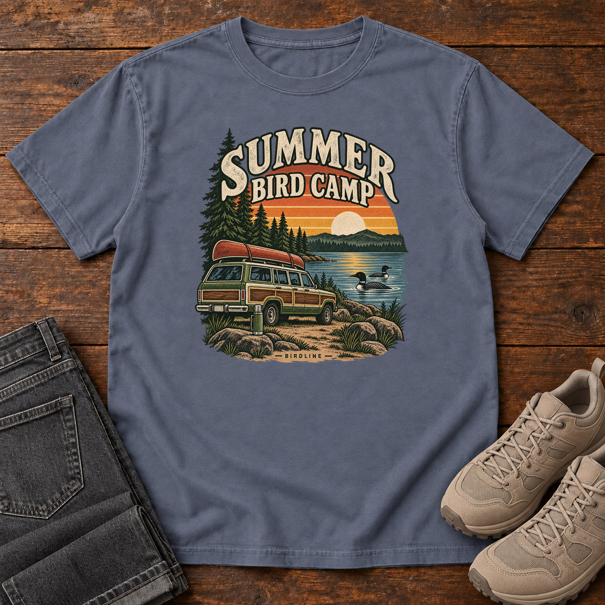 Summer Bird Camp T-Shirt in Denim, front laid flat on wood, featuring a canoe-topped vehicle beside a lake with swimming birds, pine trees and a striped sunset.