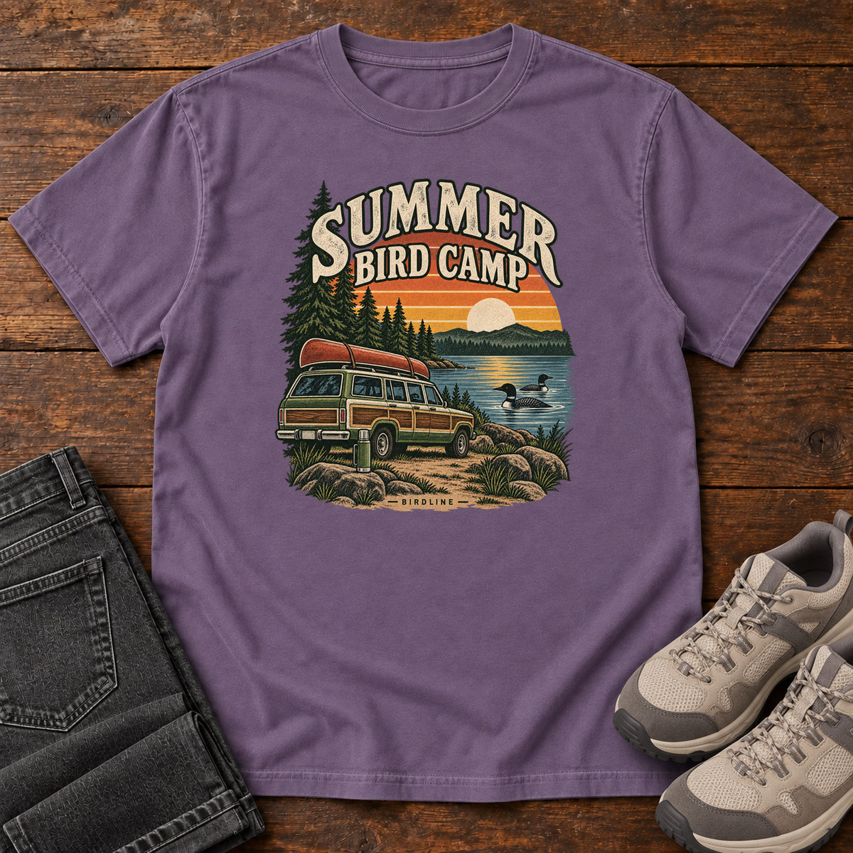 Summer Bird Camp T-Shirt in Grape, front laid flat on wood, featuring a canoe-topped vehicle beside a lake with swimming birds, pine trees and a striped sunset.